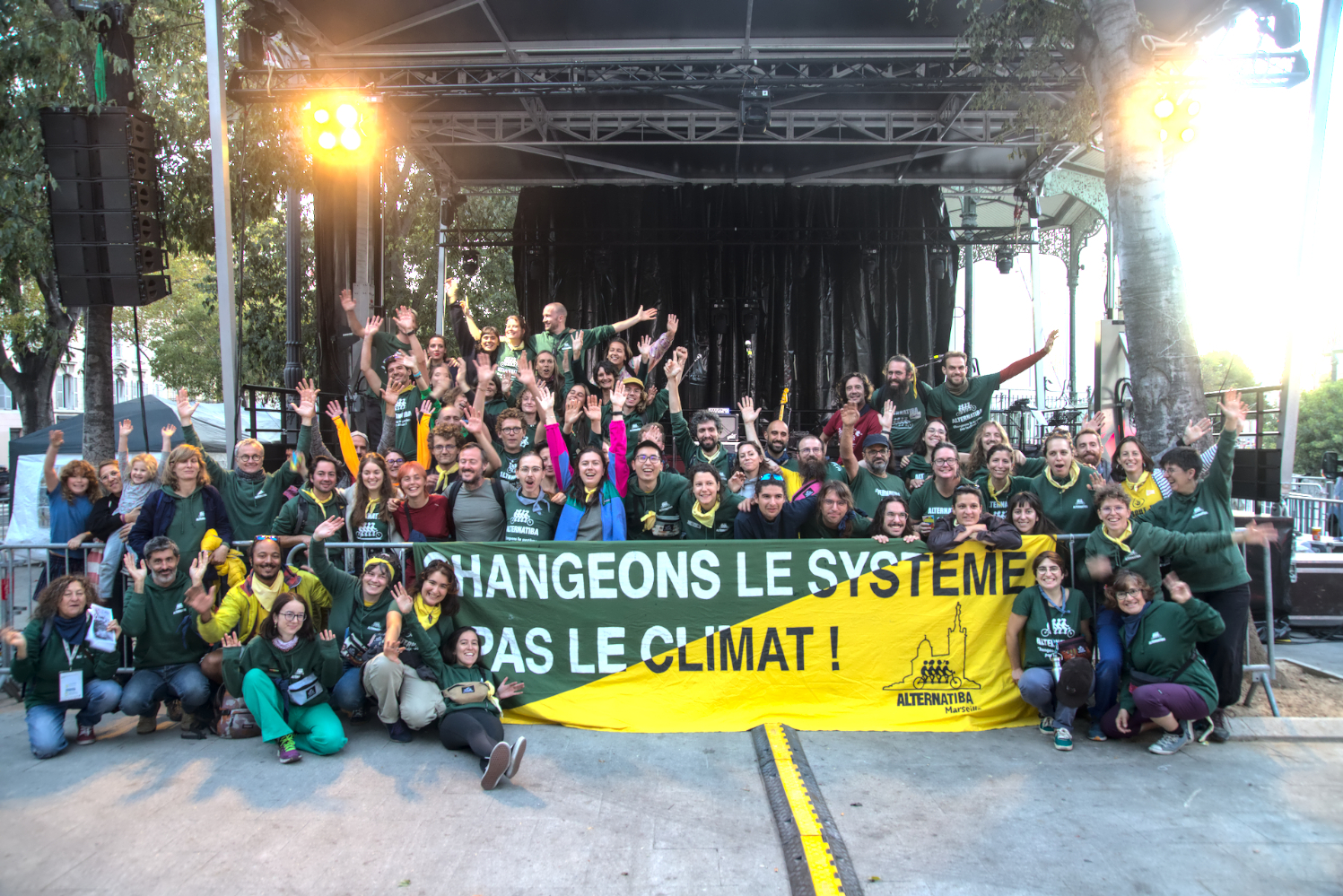 Des militant·es d'Alternatiba et ANV-COP21 sont groupé·es autour d'une banderole "Changeons le système, pas le climat". Iels lèvent les bras au ciel et sourient.